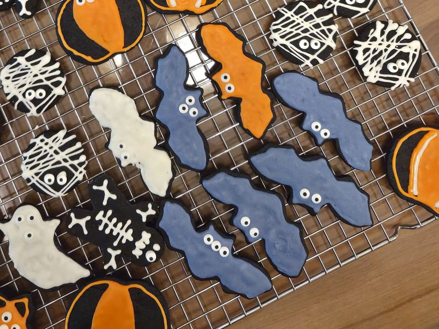 Real hand-decorated black cocoa cookies on a cooling rack: blue bats with candy eyes, white-piped mummies and a white ghost